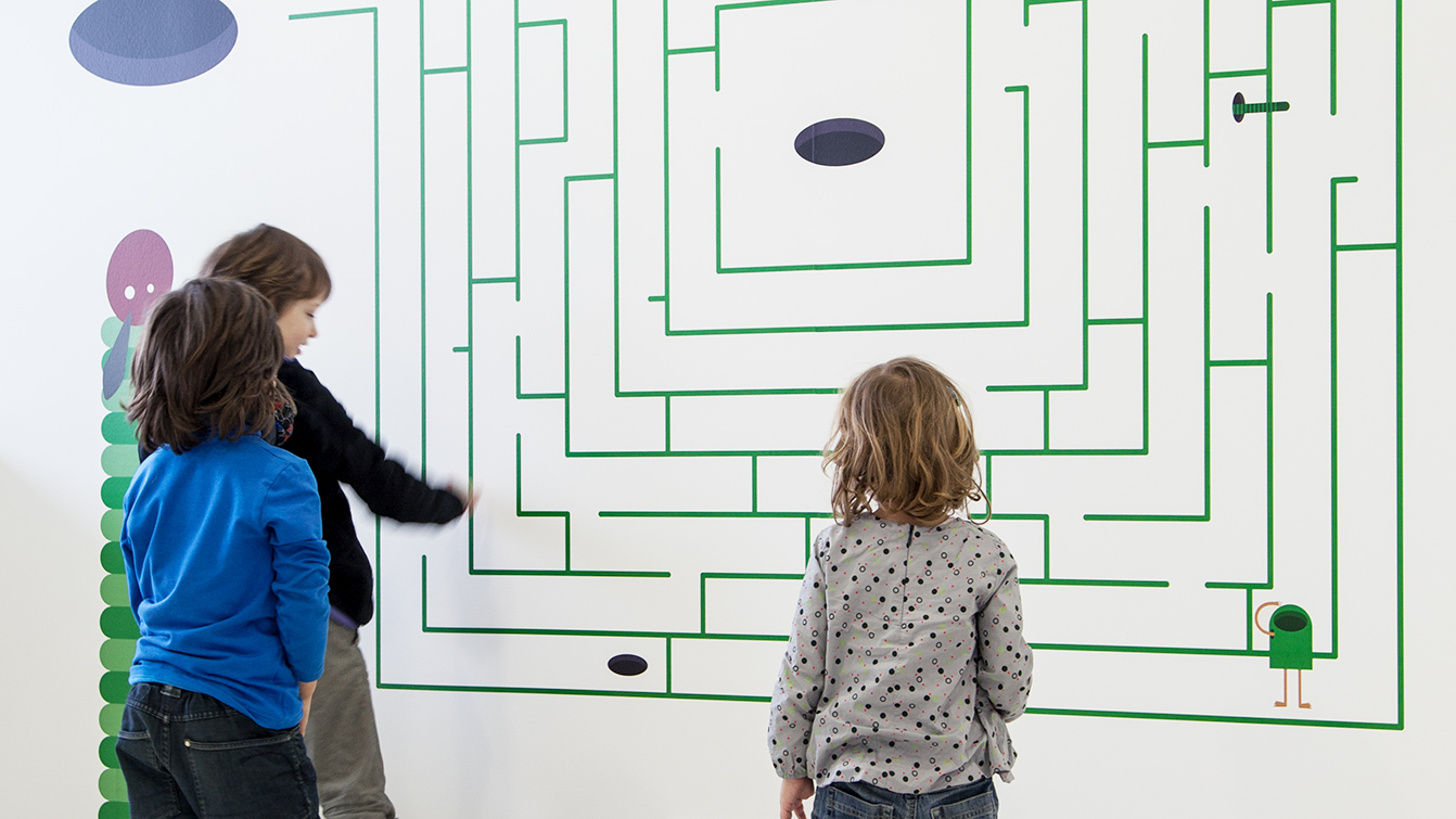 Kinder vor einer Wand stehend, auf der ein Labyrinth zu spielerischen Nachfahren mit dem Finger aufgemalt ist