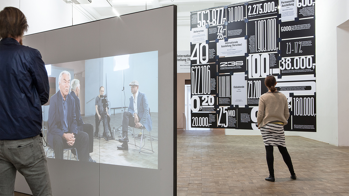 Two people standing in front of floor-to-ceiling exhibition graphic and large screen