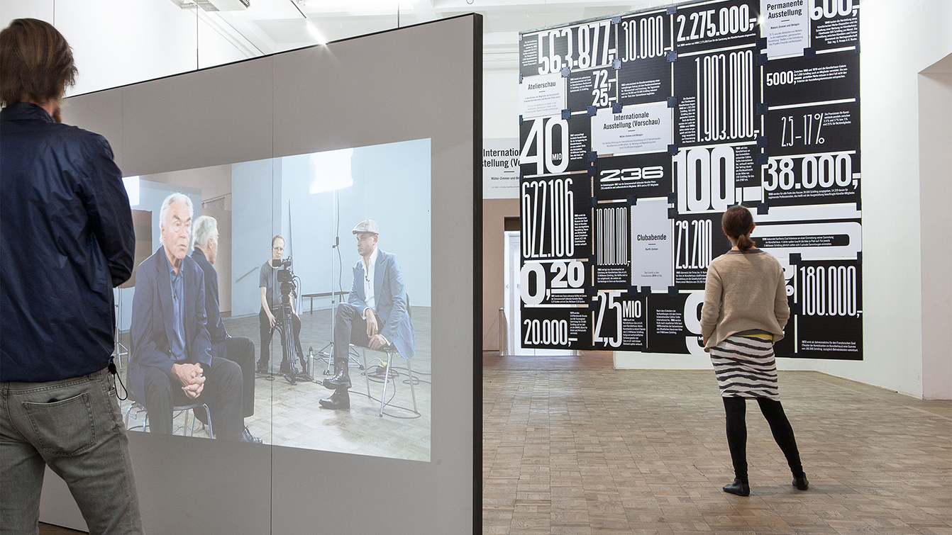 Zwei Menschen vor raumhoher Ausstellungsgrafik und einer großen Leinwand stehend