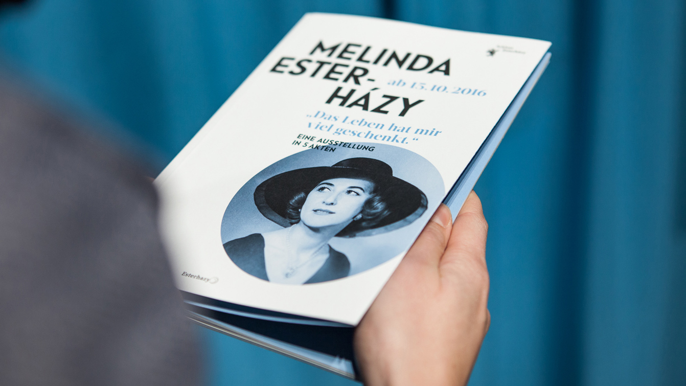 Hand die die geschlossene Ausstellungsbroschüre in der Hand hält mit Portrait der Melinda Esterhazy am Cover