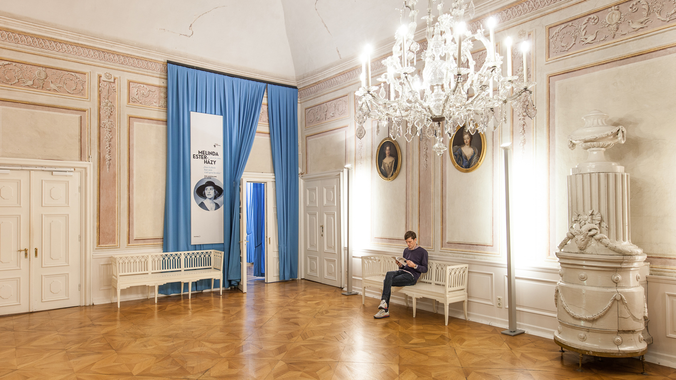 Barocker Raum im Schloss Esterhazy mit Luster an der Decke, einem Menschen auf einer Bank vor einem blauem Vorhang der zur Raumtrennung dient sitzend, mit darauf angebrachter Ausstellungsgrafik