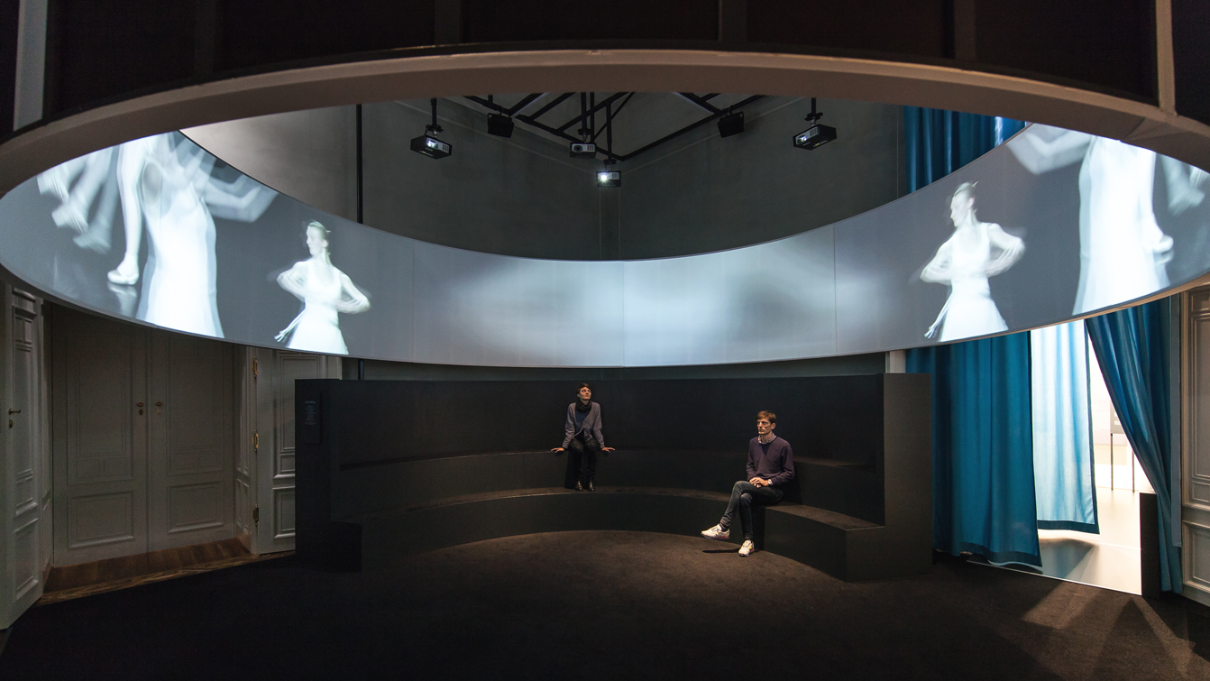 Dunkler Ausstellungsraum, mit Unteransicht der ovalen Kino-Leinwand, die als über Kopf als Streifen in der Luft hängt