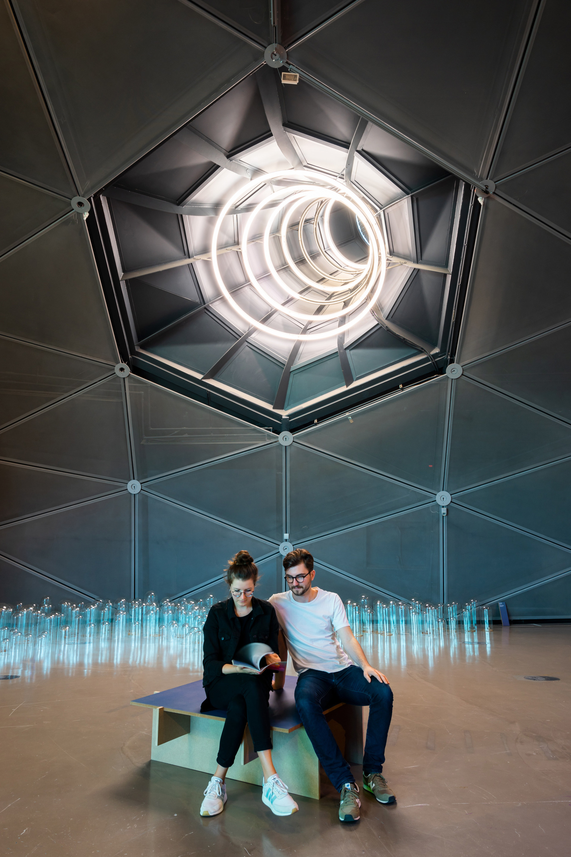 Lichtinstallation an der Decke des Kunsthauses, unter der zwei Menschen in der Mitte eines Ausstellungsraums sitzen und einen Katalog durchblättern