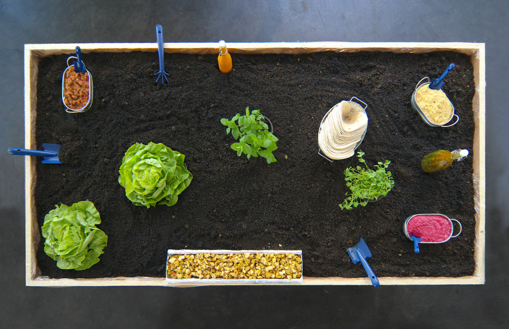Tisch von oben fotografiert, der einem Gartenbeet gleicht mit Salaten, Gartenschaufel, Blumenkisten gefüllt mit Dips, Erde aus schwarzem Couscous
