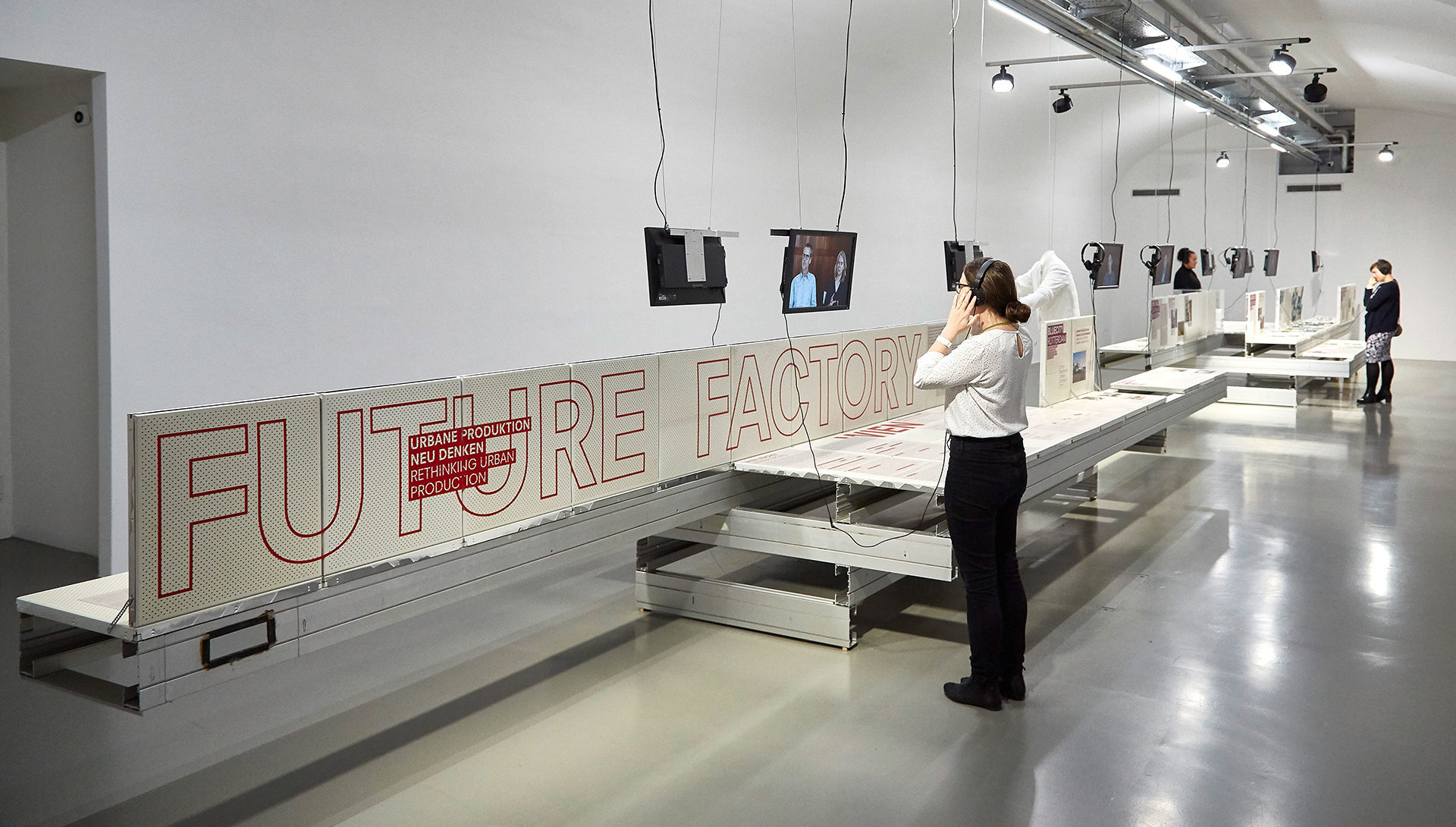 Ansicht des Ausstellungsraums der Future Factory mit Display für Projektbeispiele, Fernsehern für Interviews und Besucherinnen