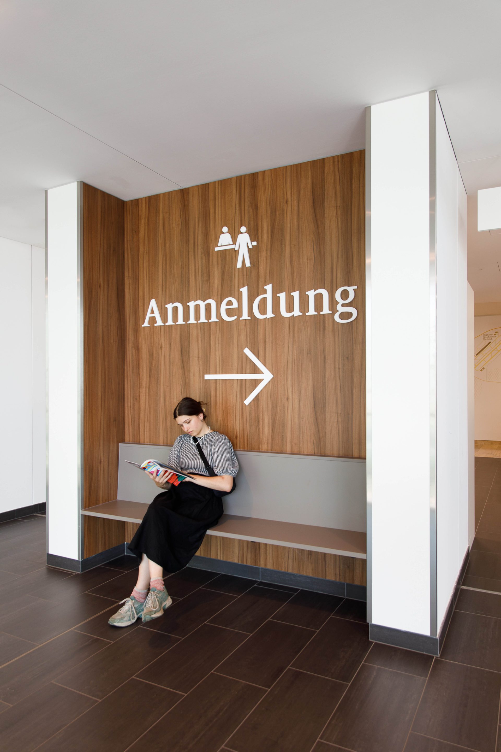 Signpost with writing and pictogram to the registration area in a seating niche where a woman is sitting with a newspaper