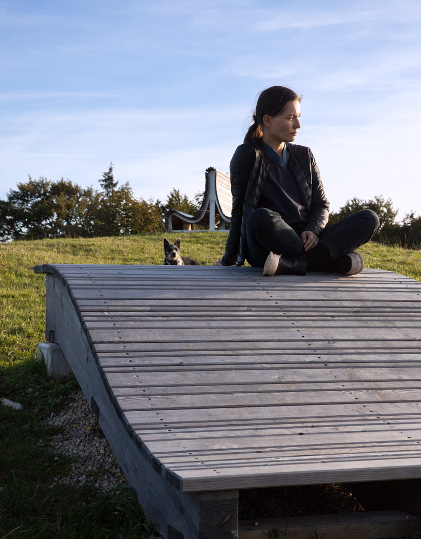 Wooden furniture in a wave shape standing on a meadow, a woman sitting on it and a dog in the background