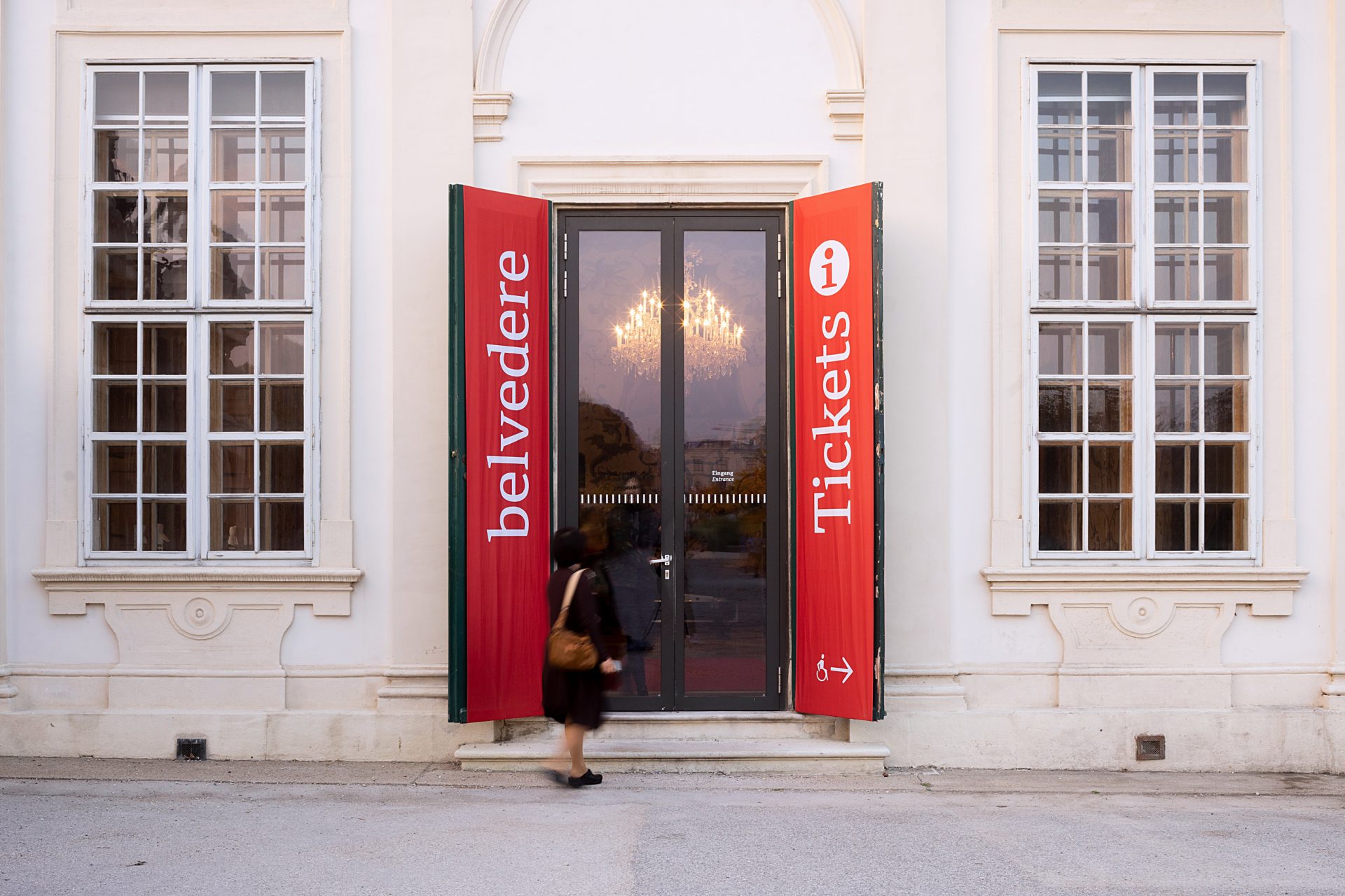 Eingangstüre aus Glas mit Seitenflügeln auf denen rote Fahnen angebracht sind, links steht Belvedere, rechts steht Tickets und Information
