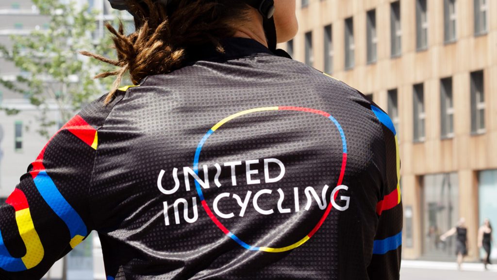 Man photographed from behind with focus on back area, wearing a black cycling dress with colorful lines and the logo of United in Cycling